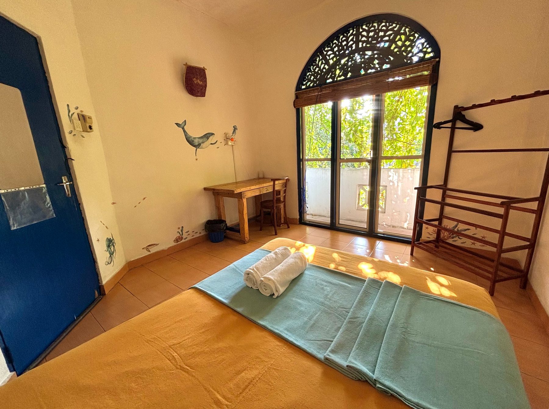 Ocean Double Room at Seconds Colombo — small hand-painted whale on the wall, deep blue door, arched colonial window, mustard floor with rolled towels